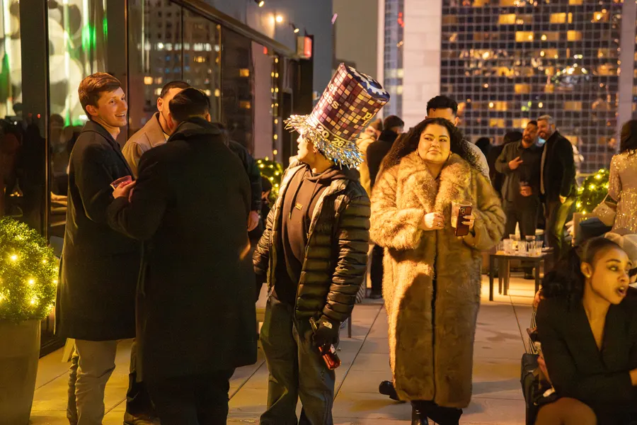 Guests on a rooftop terrace at a New Year's Eve party wearing a long faux-fur coat, a puffer jacket and wool overcoats